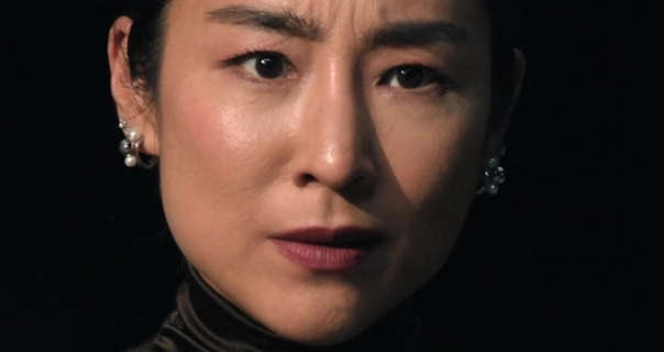 Greta Lee is seen in a dark room with a black wall behind her. She has her short black hair straightened and tucked back. Greta appears in a high neck top, mascara, pearl cluster earrings and a dark pink lipstick.