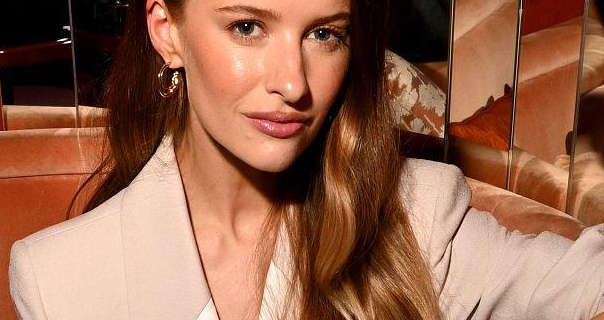 Victoria Magrath sits on a large corner sofa in front of a segmented mirror wall. She has her long light brown hair loose and in natural waves. Victoria wears a satin lapel blazer with a pair of large hoop earrings, blush and a dark pink lipstick.