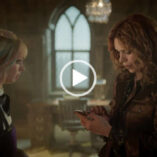 Billie Piper looks down at a small smartphone. She stands in front of Emma Myers in a large castle like dormitory room with a writing desk and ornamental window nearby. Billie has her hair styled in curls. She is seen in a long sleeve top and a brown shiny jacket.