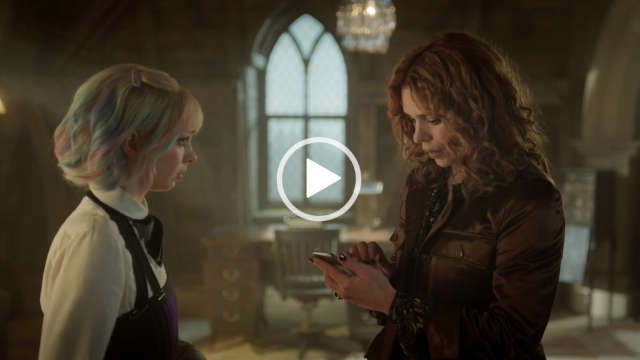 Billie Piper looks down at a small smartphone. She stands in front of Emma Myers in a large castle like dormitory room with a writing desk and ornamental window nearby. Billie has her hair styled in curls. She is seen in a long sleeve top and a brown shiny jacket.