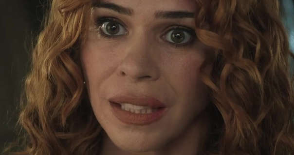 Billie Piper stands in a dark boarding school bedroom and looks a bit concerned. She wears her hair in tight curls. Billie is seen in a matt foundation, blush, black mascara and a dark pink matt lipstick.