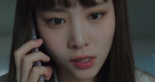 Lee Yul-eum is seen with her long black hair loose and with a fringe. She holds a silver frame smartphone to one ear. Lee Yul-eum wears foundation with mascara and a dark pink lipstick. She stands in a room at twilight in light gold sleepwear with a slightly frustrated expression as she talks.