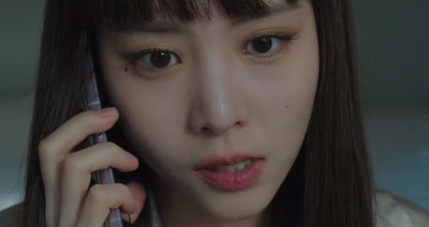 Lee Yul-eum is seen with her long black hair loose and with a fringe. She holds a silver frame smartphone to one ear. Lee Yul-eum wears foundation with mascara and a dark pink lipstick. She stands in a room at twilight in light gold sleepwear with a slightly frustrated expression as she talks.
