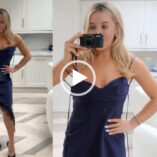 Em Sheldon appears in her white marble London kitchen in front of a white chair and white cabinets. Em is seen with her long blonde hair loose and styled naturally. She wears an asymmetrical navy strappy dress with a pair of high heel shoes.