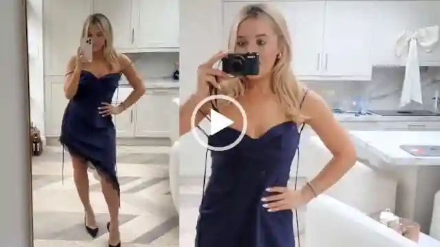 Em Sheldon appears in her white marble London kitchen in front of a white chair and white cabinets. Em is seen with her long blonde hair loose and styled naturally. She wears an asymmetrical navy strappy dress with a pair of high heel shoes.
