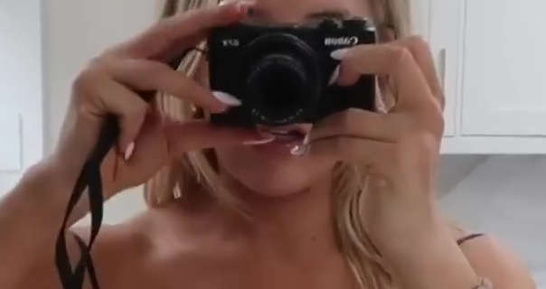 Em Sheldon holds her black Canon vlogging camera in front of her face. She stands in front of her white kitchen cabinets and cream marble splashbacks. Em is seen with her long black hair loose and styled naturally. She wears a pink gloss lipstick and long white gel nails with a navy strappy dress.