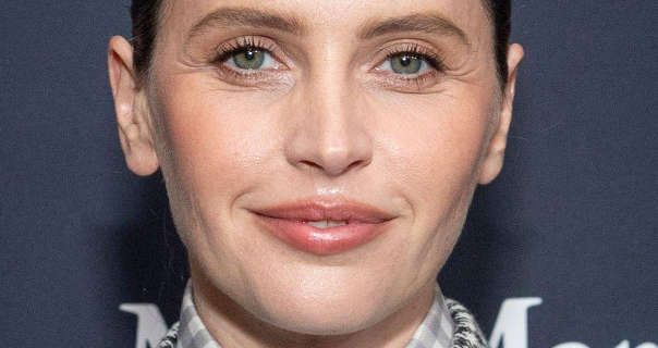 Felicity Jones appears in front of a dark grey wall that has white writing across it. She wears her black hair tied back and in a bun. Felicity is seen in a matt foundation, light blush, mascara and a dark pink gloss lipstick. She wears chequered clothing and no jewellery.