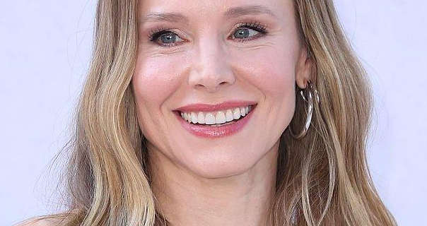 Kristen Bell smiles as she stands in front of a dark white wall. She wears her long blonde hair loose and slightly curled. Kristen is seen in a pink gloss lipstick, black mascara and a pair of large gold hoop earrings.