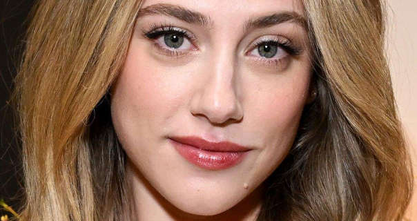 Lili Reinhart is seen close up with her dark blonde hair loose and a little wavy. She appears in front of a black and cream wall. Lili wears a matt full coverage foundation with soft blush, mascara and a dark red gloss lipstick.