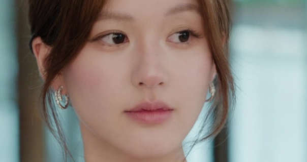 Lusi Zhao glances to one side. She stands in front of a row of large windows with daylight shining through. Lusi has her light brown hair tied up with a few strands down around her face. She wears a light pink blush with a pair of medium size hoop earrings and a light pink lipstick.