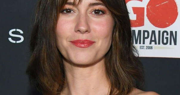 Mary Elizabeth Winstead appears in a light foundation, pink blush, black mascara and a dark red gloss lipstick. She stands in front of a dark grey poster wall with white text and a white, red and dark grey logo across it. Mary has her long brown hair loose and in soft waves.