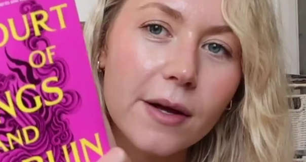 Meg Says holds up a pink book that has yellow writing and a and a swirling sword design. Meg has her blonde hair tousled and loose. She sits in front of a cream wall and stacked wicker storage baskets. Meg appears in soft foundation and blush with a pair of small gold earrings and a dark pink matt lipstick.