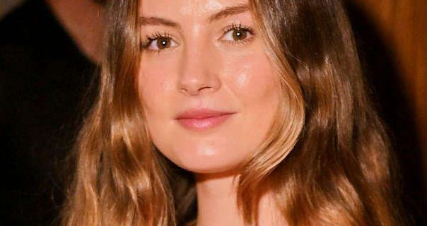 Niomi Smart is seen in a dark room at an event. She wears her light brown hair loose, styled naturally and with blonde colouring. Niomi smiles as she looks into the camera. She wears a glowy foundation with a light pink sheen lipstick and black lash curling mascara.