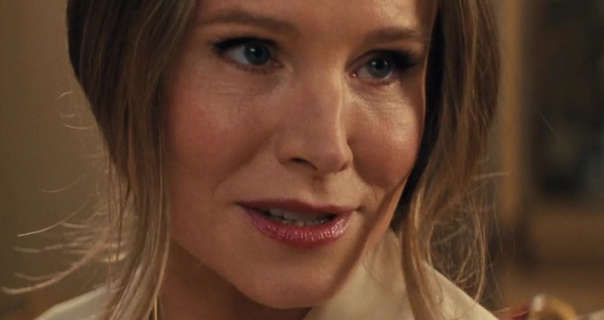 Kristen Bell sits in a beige dining room area with a dark gold framed painting on the wall behind her. Kristen has her long dark blonde hair tied back with strands pulled down to frame her face. She wears a dewy natural foundation with a red blush and a dark red lipstick.