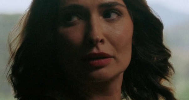 Karolina Wydra stands in front of a large glass window during the day with green trees and shrubs outside. She wears her hair curled and loose around her shoulders. Karolina is seen in a pink lipstick and light blush. She glances to one side with her face partially in shadow.