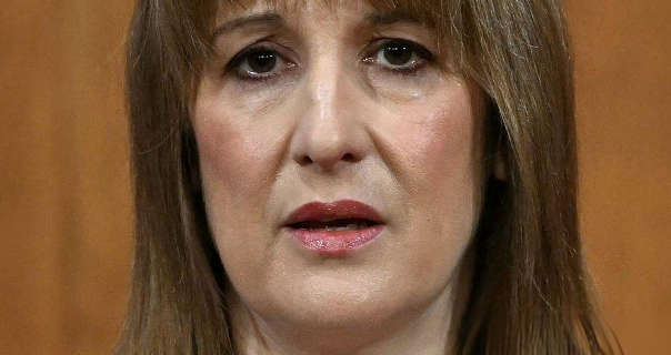 Rachel Reeves stands in front of a wood panel wall. She is seen with her light brown hair down and loose. Rachel wears a pink blush with mascara and a dark pink lipstick. She looks straight ahead with a concerned and serious expression.