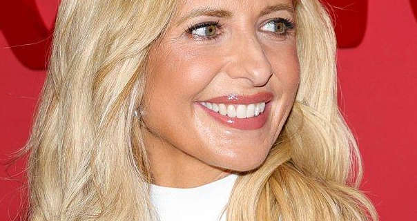 Sarah Michelle Gellar has her long blonde hair down and styled in waves. She appears in front of a pastel red wall with darker red writing across it. Sarah is seen in a dark pink gloss lipstick, dewy foundation, black mascara and a white top. She smiles and looks off to one side.