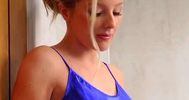 Serenity leans against a wood slat doorway in a cream splashback tiled bathroom. She wears a bright blue strappy nightdress with her long blonde hair tied up and back. Serenity appears in a pink gloss lipstick, foundation and eyelash extensions.