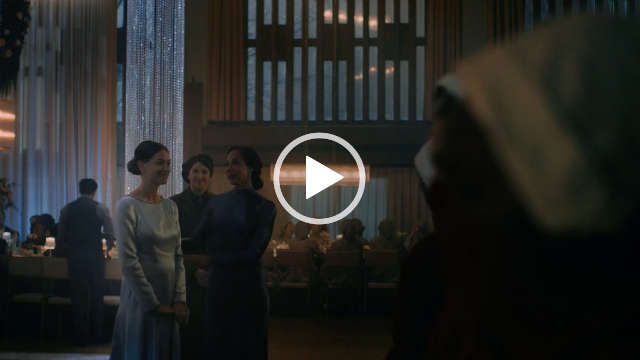 Yvonne Strahovski stands in a large dining room with a long table of seated people behind her. Yvonne speaks to a gathered crowd. She wears a long sleeve light blue gown with her long blonde hair tied back in a bun.