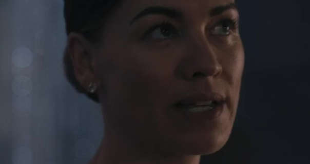 Yvonne Strahovski glances upwards while standing in a dark reception room. She wears her blonde hair tied back in a bun. Yvonne stands in front of a grey wall and white sheer curtains. She is seen in a matt dark pink lipstick, foundation, mascara and a pair of large diamond earrings.