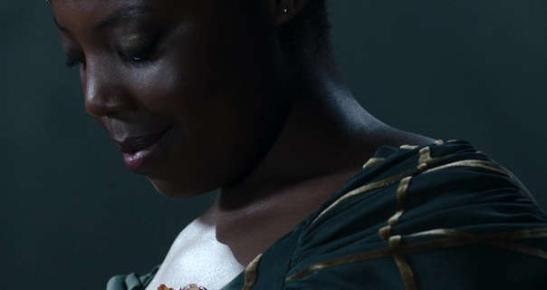 Mimî M Khayisa wears a dark blue cap that has a gold threaded pattern. She has her black hair tied up in a ornate bun. Mimî stands in a dark stone room. She is seen in black mascara, metal earrings and a matt lipstick.