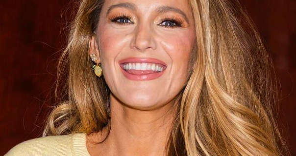 Blake Lively is seen in front of a dark wood wall. She wears her long hair loose, styled and with streak highlights. Blake appears in a pale yellow thin knitted top, gold earrings, foundation, blush, mascara and a pink gloss lipstick.