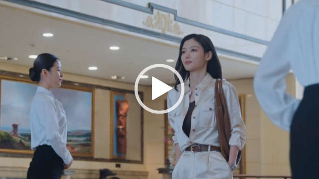 Kim Yoo-jung walks through a large open plant room with cream tiles and paintings on the walls. She passes assistants in formal attire. Kim Yoo-jung has her long black hair loose and pushed back. She wears a light gold unbuttoned shirt over a black top and gold trousers. She carries a large brown bag over one shoulder and is seen in full coverage foundation and a matt light pink lipstick.