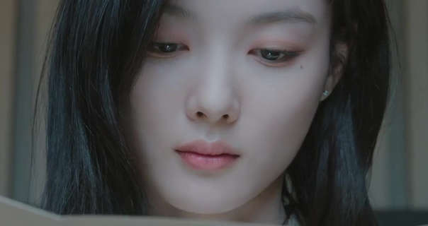 Kim Yoo-jung appears with her long black hair loose and naturally straight. She wears a matt foundation with pink eye shadow and a light pink lipstick. Kim Yoo-jung sits in front of a vertical slat effect wall. She looks down at a large cream wedding invitation.