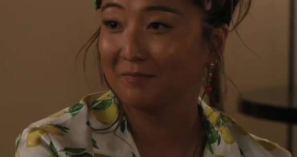 Ashley Park smiles a little as she appears in a small sitting room. She wears her thick black hair pulled up in a messy bun. Ashley is seen in a white lemon print pyjama tip, hanging earrings, dewy foundation and a dark pink lipstick.