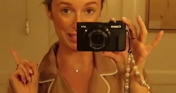 Josie Irons holds a black Canon vlog camera in one hand and points up and to one side with her other hand. She stands in front of a cream wood panel wall at her Cotswolds home. Josie wears her hair tied up in a bun. She is seen in a fresh foundation, mascara and a dark pink matt lipstick.