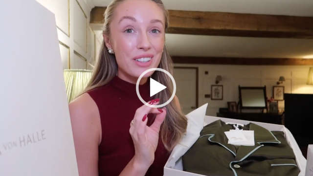 Josie Irons holds an open white Olivia Von Halle box with a pair of deep green turquoise trim pyjamas. Josie has her long hair loose and tied back. She wears a sleeveless top with foundation, matt lipstick and a pair of pearl earrings. Josie sits in a corner of a white wood and exposed natural beam bedroom in her Cotswolds home.