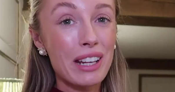 Josie Irons sits in the corner of a large bedroom at her country home. She is seen in front of a low wood beam ceiling, white panel walls and a green lamp. Josie wears her dark blonde hair loose and pinned back. She appears in a pair of pearl earrings, matt foundation, pink blush, mascara and a pink matt lipstick.