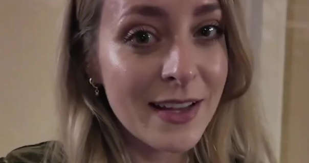 Fleur De Force is seen with her long blonde hair loose and styled naturally. She stands in front of a light beige wall and a white wooden door frame in her home. She wears a dewy foundation with black mascara, a dark pink matt lipstick and delicate silver earrings.