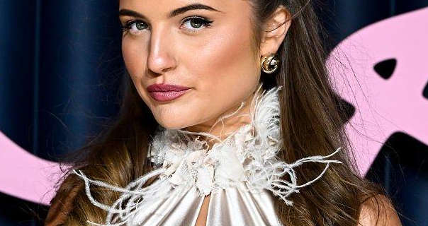 Grace Foley stands in front of a dark navy wall that has light pink text across it. She is seen with her long brown hair loose and pinned back. Grace wears a flowing gold feather trim gown. She pairs gold earrings with a dark pink lipstick, black mascara, matt foundation and a defining blush.