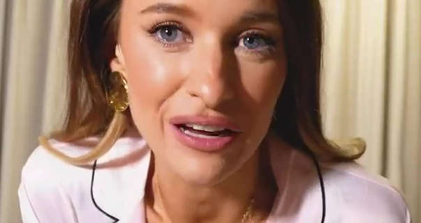 Victoria Magrath leans into the camera. She has her hair loose and pushed back and with blonde highlights. Victoria is seen in a light pink and black trim pyjama top, standing in front of drawn cream bathroom curtains. She appears in a pair of large gold sheet circular earrings, foundation, blush, mascara and a dark pink matt lipstick.