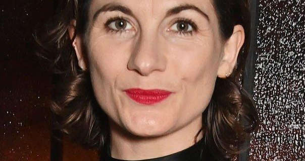 Jodie Whittaker smiles and looks in the camera. She wears her dark brown hair cut short and curled. Jodie is seen wearing a bright red lipstick, matt foundation and black mascara. She stands in front of a black and brown shimmery wall.