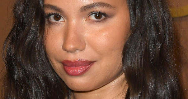 Jurnee Smollett is seen with her thick long black hair loose and in soft waves. Jurnee wears blush with black mascara and a dark red lipstick. She stands in front of a deep gold poster wall.