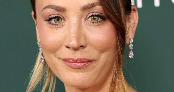 Kaley Cuoco stands in front of a dark forest green wall that has some white text on it. She wears her blonde hair tied up and with end highlights. Kaley is seen in a matt foundation, mascara, pink gloss lipstick and a pair of large tiered diamond earrings.