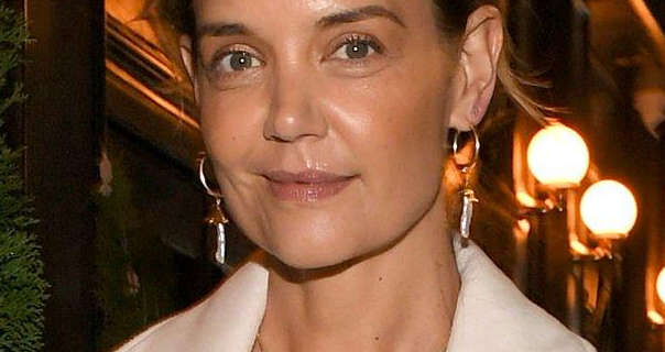 Katie Holmes is seen at night in front of a row of globe lights that case an orange glow. She appears with her light brown hair tied back. Katie wears a pair of hanging gold gemstone earrings with a long cream coat, foundation, black mascara and a dark pink lipstick.