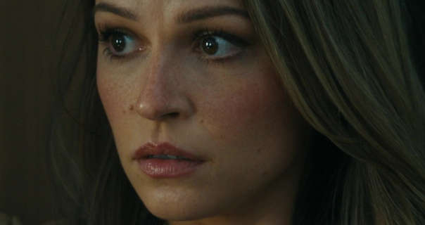 Kayla Wallace looks to one side with a puzzled and slightly concerned expression. She is seen against a dark brown backdrop with her hair loose and in gentle waves. Kayla wears a dark pink gloss lipstick with blush highlights and mascara.