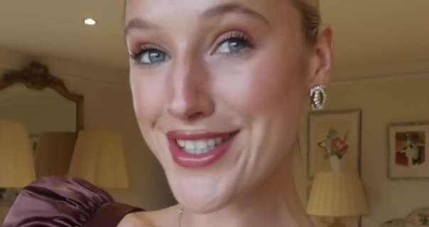 Leonora Smee stands in her large cream and white dressing room at home. She has her long blonde hair tied up and back in a tight bun. Leonora wears a dark pink gloss lipstick with black mascara, foundation, blush and gold jewellery.