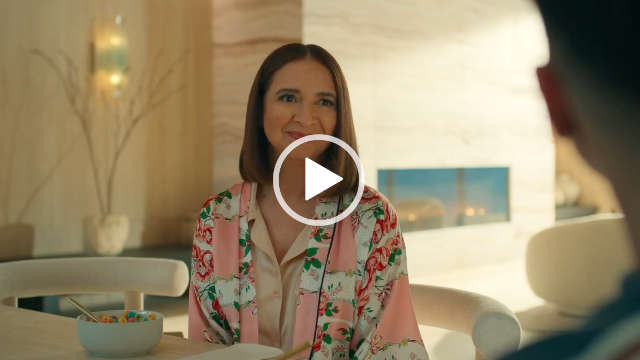 Maya Rudolph sits at a large light wood table and on a white wooden chair. She appears in a large open plan kitchen dining area. Maya has her brown hair loose and cut to shoulder length. She wears a floral light pink robe over a pair of pink pyjamas.