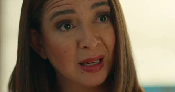 Maya Rudolph is seen in front of a large window in a large open plan living room as the sun is just starting to rise. She wears her thick brown hair loose and straightened. Maya stars in blush, mascara and a dark red lipstick.