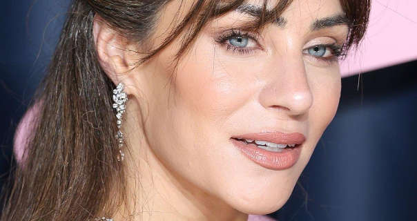 Lydia Millen is seen in front of a dark grey and light pink event wall. She wears her long brown hair pinned back and with a long fringe. Lydia wears a pair of dangling diamond earrings with a matt foundation, dark blush, black mascara and a dark pink satin lipstick.