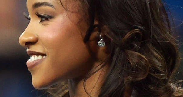 A Detroit Lions cheerleader is seen in profile. She has her thick black hair loose and in messy curls. The cheerleader appears in a soft blush, dark mauve lipstick, heavy mascara and a pair of small silver gemstone earrings.