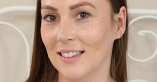 Harper is seen in front of a beige wall and a white thin wire frame bed headboard. She has her light brown hair loose and straightened. Harper appears in black mascara, a light pink lipstick and blush with no foundation.