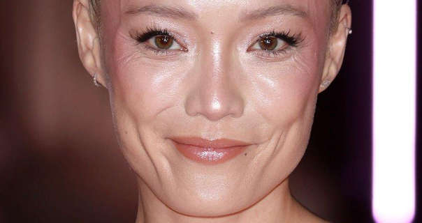 Pom Klementieff is seen with her hair tied back in a bun. She wears light foundation with a soft blush, dark pink sheen lipstick and black mascara. Pom stands in a black and neon pink room.