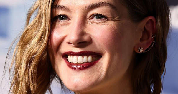 Rosamund Pike is seen with blonde highlights through her shoulder length wavy hair. She appears in front of a blue and white film poster wall. Rosamund wears a matt foundation with blue, mascara, dark red lipstick and a few small silver earrings.