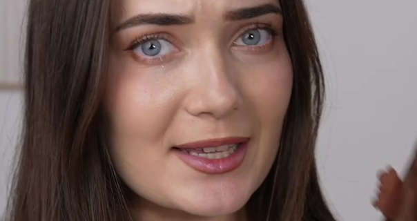 Roxxsaurus is sits in front of a dark white wall. She has her long brown hair loose and straightened. She wears a matt foundation with curled lashes and a dark pink lined lipstick. She holds a brown Shein ball gown just in frame.