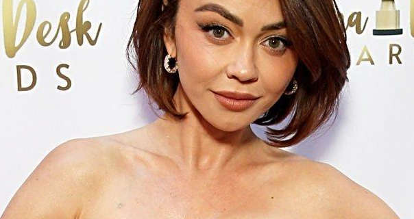 Sarah Hyland stands in front of a dark white wall that has shiny gold text and logos on it. She wears her hair cut to shoulder length and volumised. Sarah is seen in a strapless dress, dark pink lipstick, mascara and a pair of gem studded gold earrings.